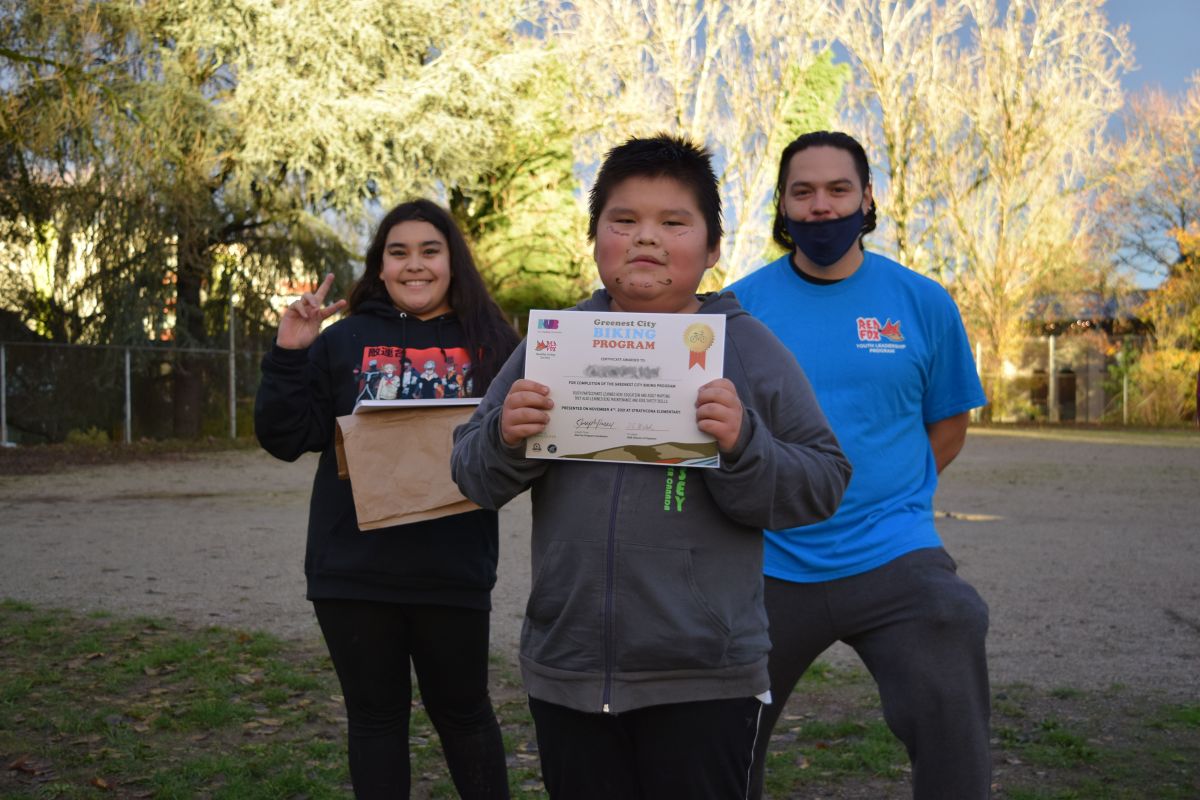 A student from the Greenest City Biking Program holds his program completion certificate. Another student and program coordinator stand in the background behind him.