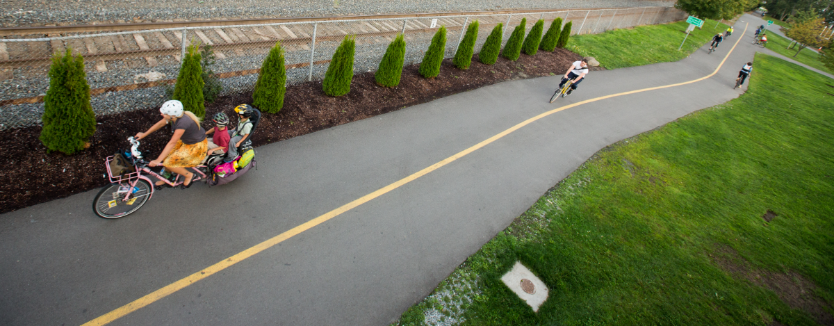 An aspirational photo of a cycle highway that runs parallel to a railroad track separated by a fence. A young woman rides a cargo bike with her two children on the back. We see several other people cycling in both directions along the cycle highway behind her.