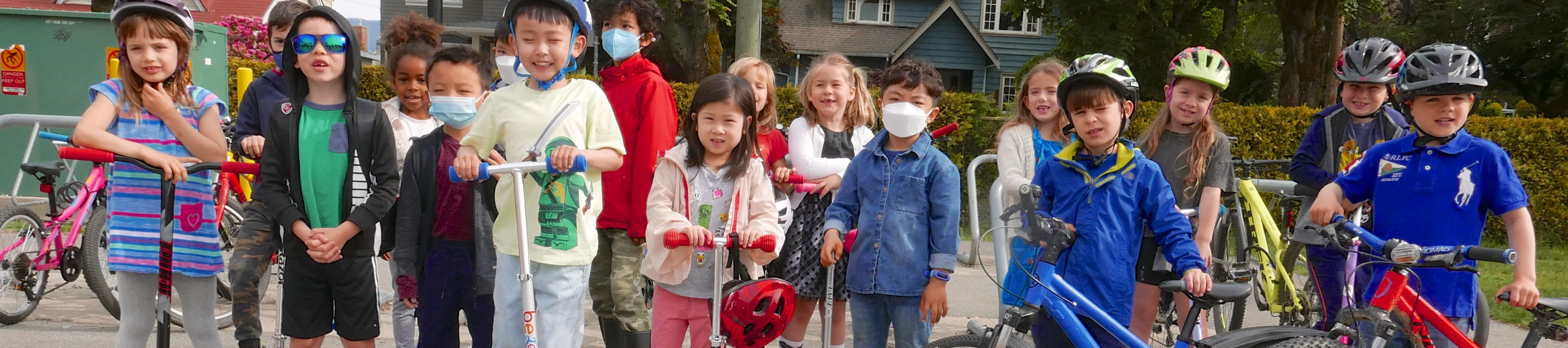 A group photo of elementary school-aged children roughly 7 years of age standing with their bikes and scooters on school grounds.