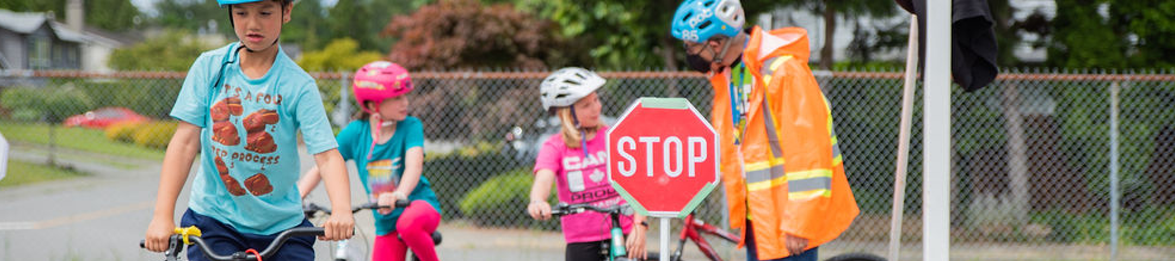 Three students about 11 years old ride their bikes on school grounds. A bike education instructor is talking to one of the students. 