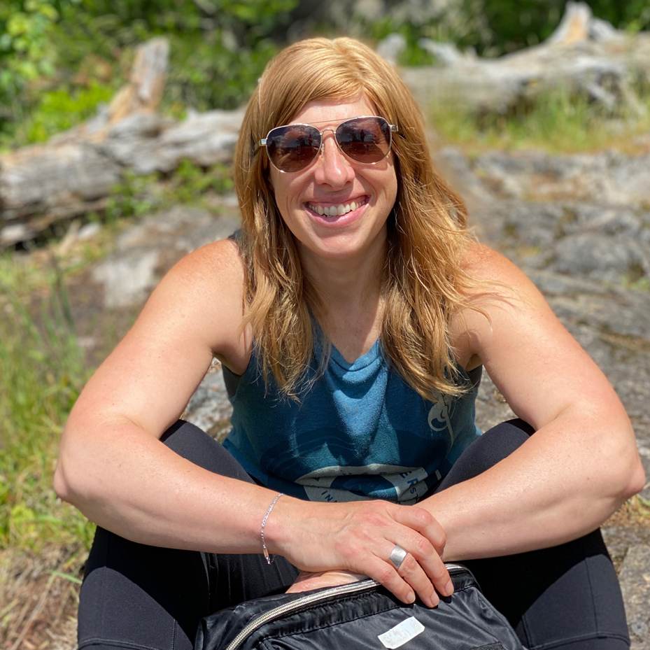 A photo of Alexandra Flynn - a woman in her 40s with long red hair. She is wearing sunglasses and a blue tank top.