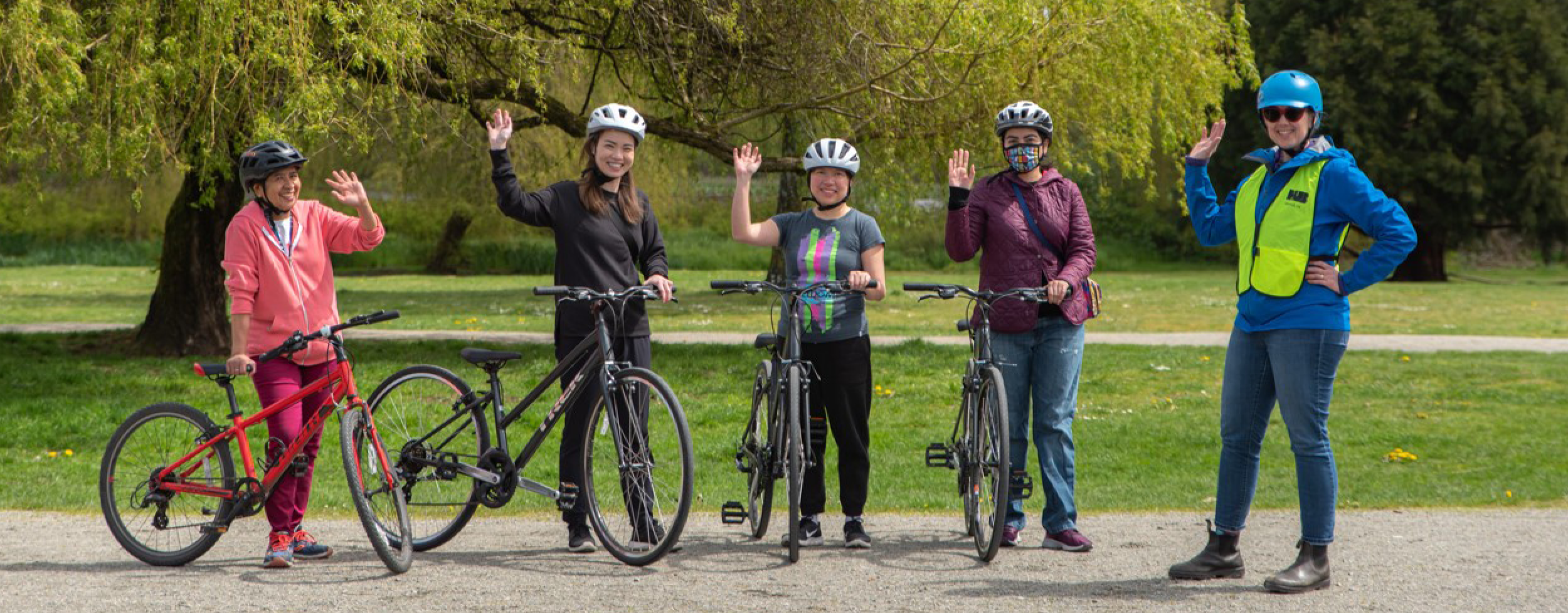 Five adult women stand in a row on a gravel field and wave at the camera. Four of these women have bikes and are taking a HUB Cycling course. The other woman is the bike education instructor.