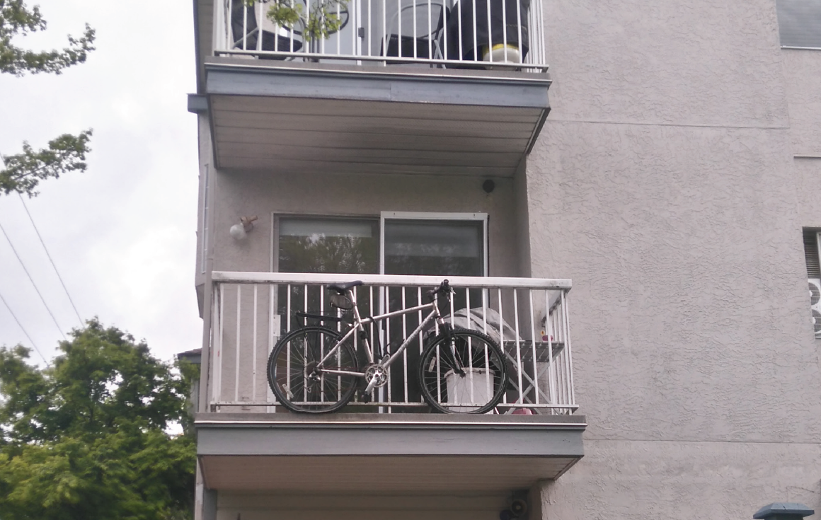 A bike sits outside on a second floor balcony of an older apartment building.