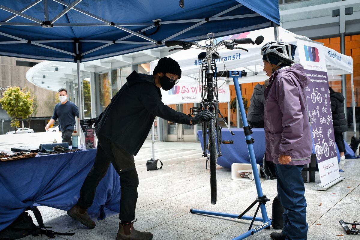 A bike mechanic spins the wheel of a participant's bike at a Knowledge HUB station. The participant, an Asian woman in her 50s or 60s, stands and watches.