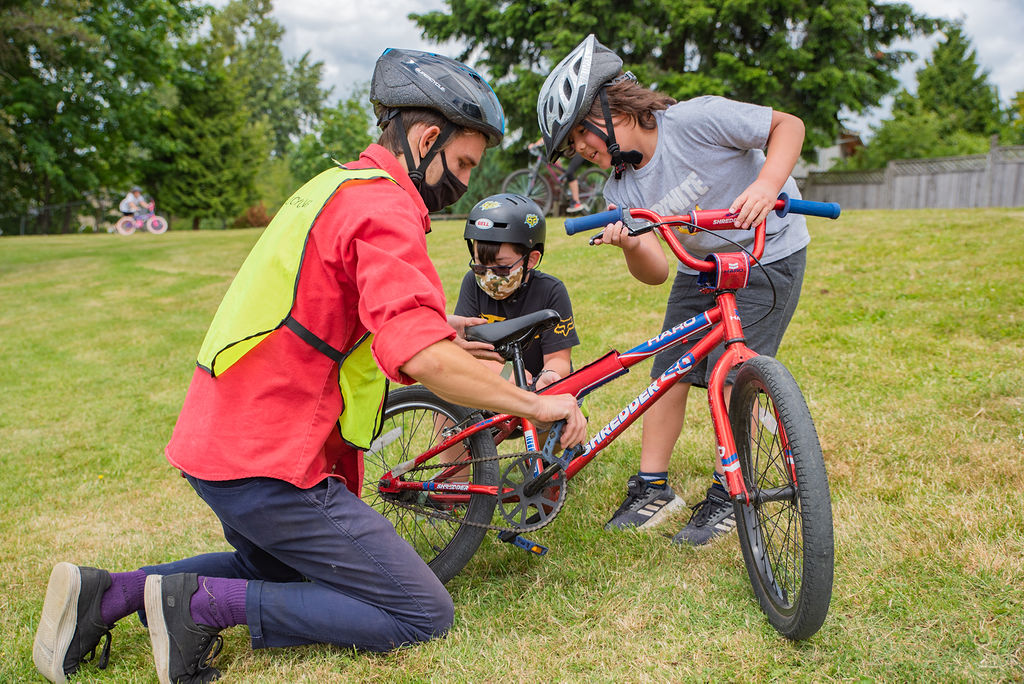 A bike education instructor kneels on the ground beside a bike as he turns the pedal with his hands. Two young buys watch him and learn.