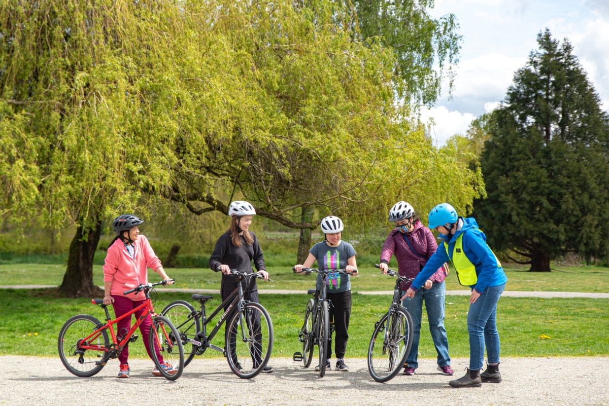 A photo taken at a Newcomer Bike Mentorship Program course. A bike education instructor points to the wheel of a participant's bike. All four, female-presenting students watch and learn.