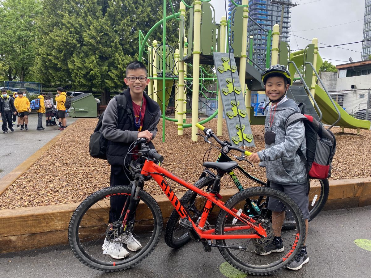 Two boys about 11 years of age smile for the camera as they stand beside their bikes in front of a school playground.