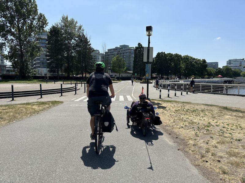 The image shows two cyclists on a designated shared-use path next to a waterfront. One cyclist is riding a traditional bike, while the other uses a recumbent trike with a safety flag. The path is bordered by green spaces, trees, and buildings on one side, and a wooden boardwalk along the water on the other. The scene is sunny with clear skies.